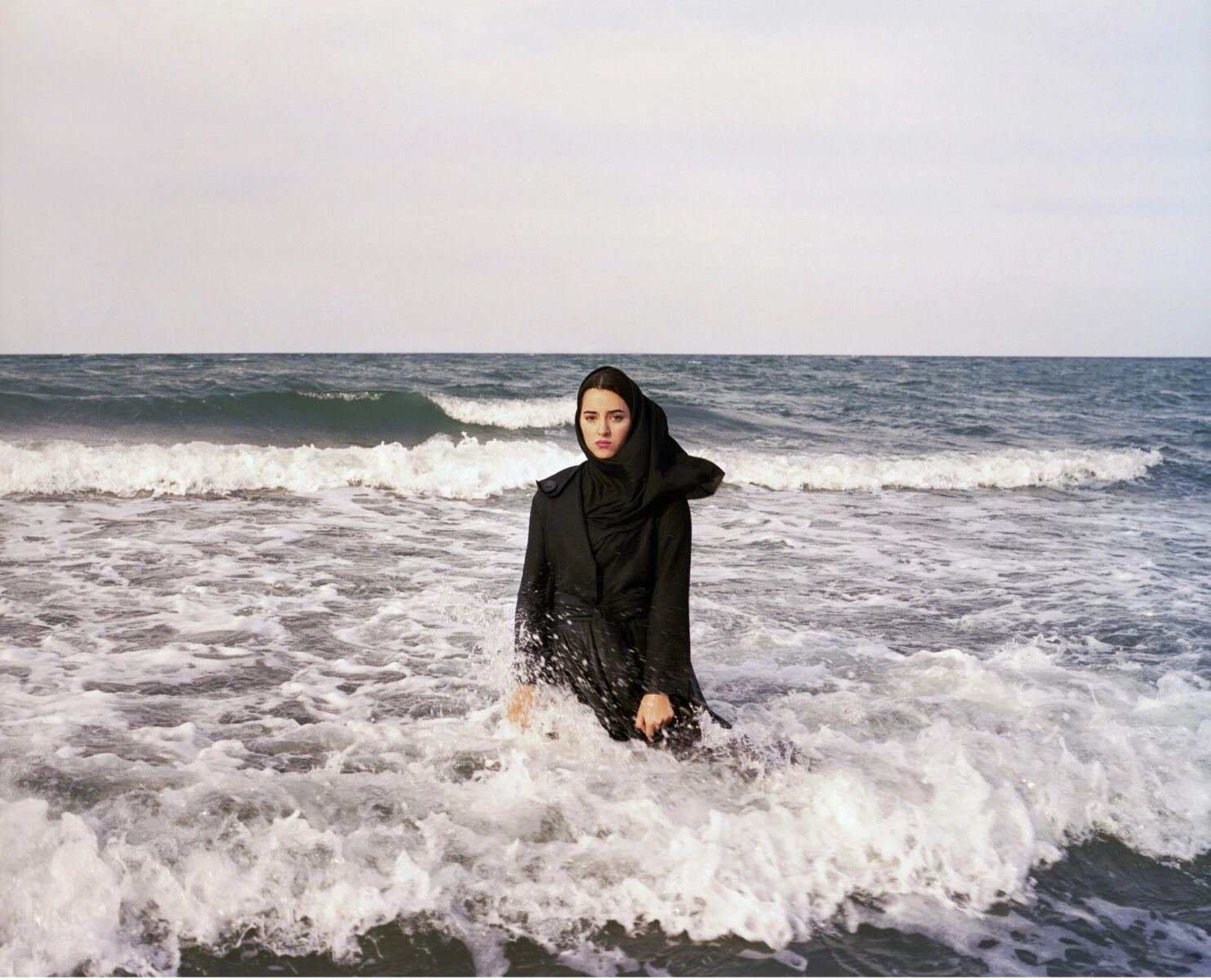 A woman, Sahar, stands clothed in a black coat and black hijab center of frame, looking directly at the camera. She is surrounded by waves up past her knees. The water is blue with frothy white caps and the sky is gray.